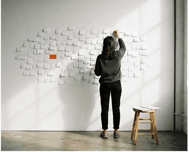 Uma pessoa a colocar um post-it numa parede coberta de notas brancas, com uma nota laranja entre elas